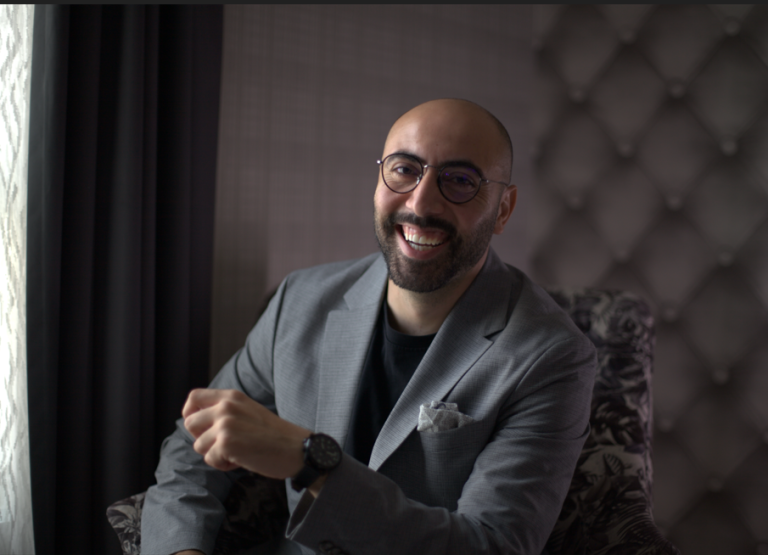 A smiling man with a bald head, glasses, and a beard, wearing a gray suit and black shirt, sits in a patterned chair near a window with soft light coming through. The background is softly lit with a tufted wall.