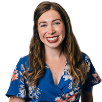 A woman with long wavy brown hair, wearing a blue floral dress, smiles at the camera against a plain white background.