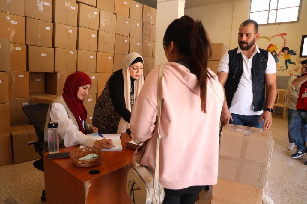 Several people stand at a desk in a room filled with stacked cardboard boxes. Demonstrating local humanitarian leadership in the Middle East, two women behind the desk write and interact with people, while a man stands nearby observing.