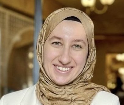 A woman wearing a tan patterned hijab and a white blazer smiles at the camera in a warmly lit indoor setting with soft lights and blurred background.