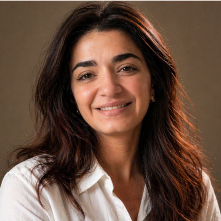 A woman with long, dark, wavy hair and light skin smiles gently at the camera. She is wearing a white shirt and gold earrings, posing in front of a plain brown background.