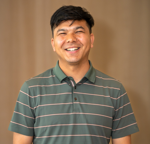 A man with short dark hair smiles broadly while wearing a green, collared, short-sleeved shirt with thin horizontal stripes. The background is plain and softly blurred.