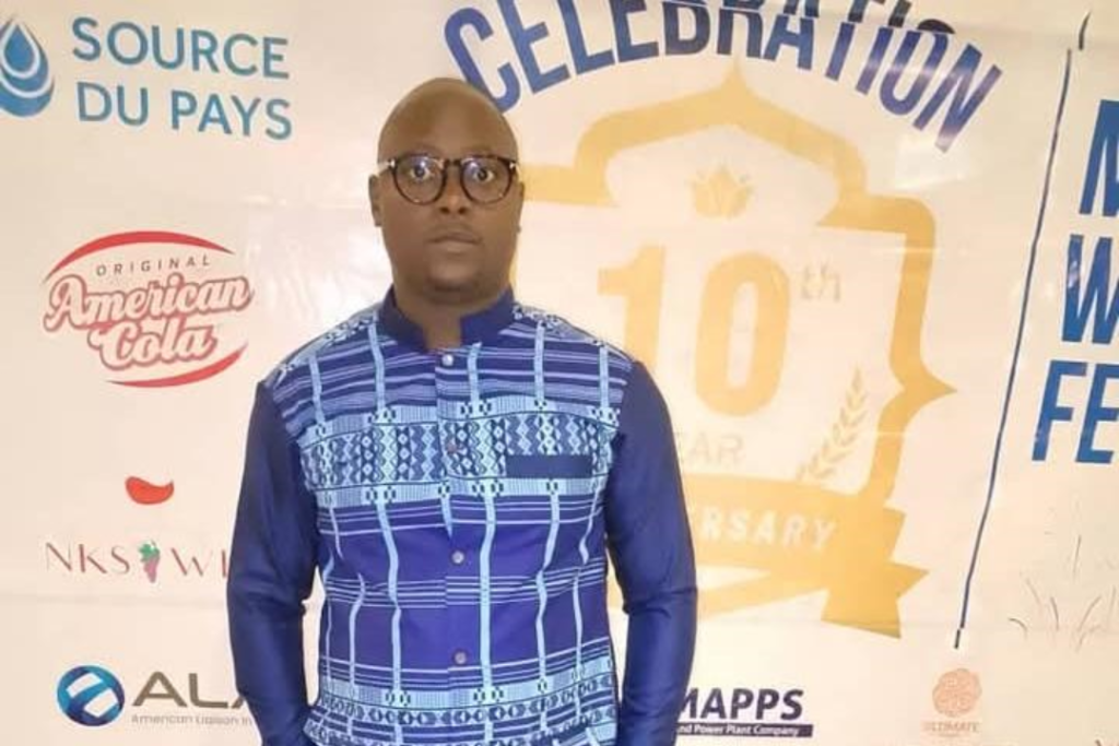 A man wearing glasses and a blue patterned shirt stands in front of a backdrop featuring various logos and text celebrating a 10th anniversary event.