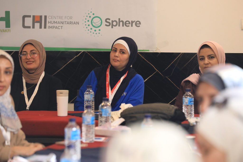 Three women in hijabs sit at a table with water bottles and notepads, attending an indoor conference—a milestone for humanitarians in Gaza. A banner behind them displays the logos for Centre for Humanitarian Impact and Sphere.