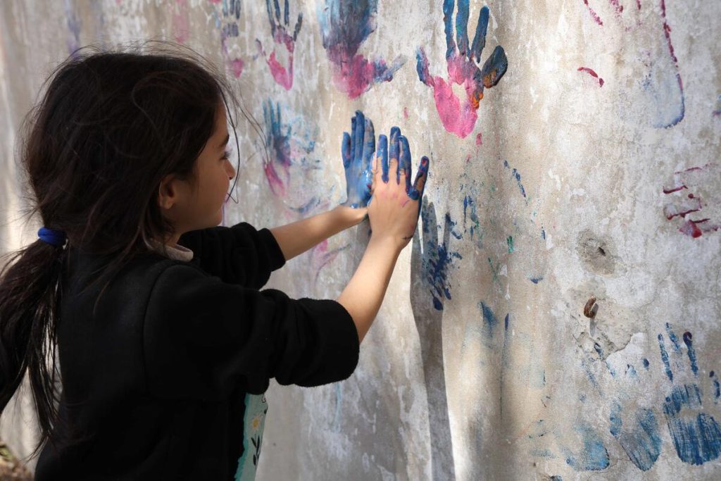 A young girl presses her painted blue hands onto a wall covered with colorful handprints, creating art outdoors.