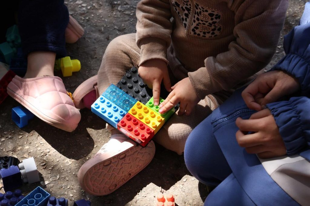 Three children sit on the ground, playing with colorful plastic building blocks. Only their hands and legs are visible, capturing a moment of hope and unity that resonates with the spirit of World Refugee Day 2026.