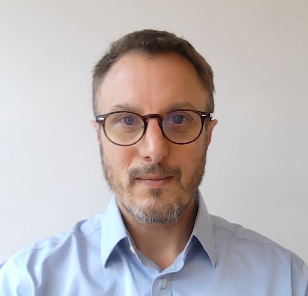 A man with short brown hair, a trimmed beard, and glasses is wearing a light blue collared shirt and looking directly at the camera against a plain white background.