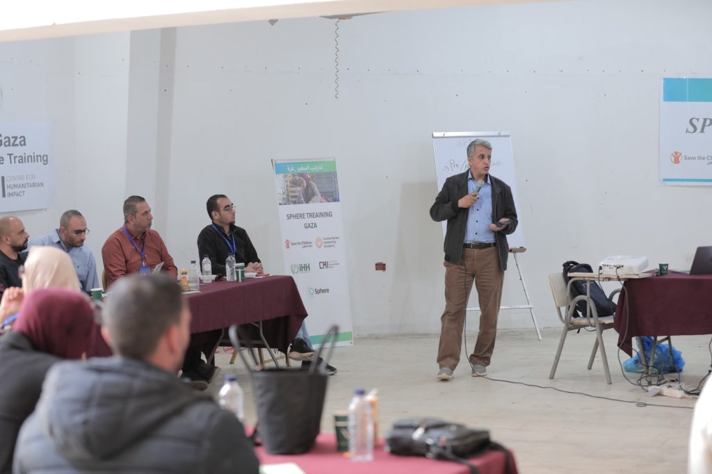 A man stands and speaks with a microphone in front of seated participants at a training session—a milestone for humanitarians in Gaza. Banners, a projector, and presentation materials are visible in the bright, indoor room.