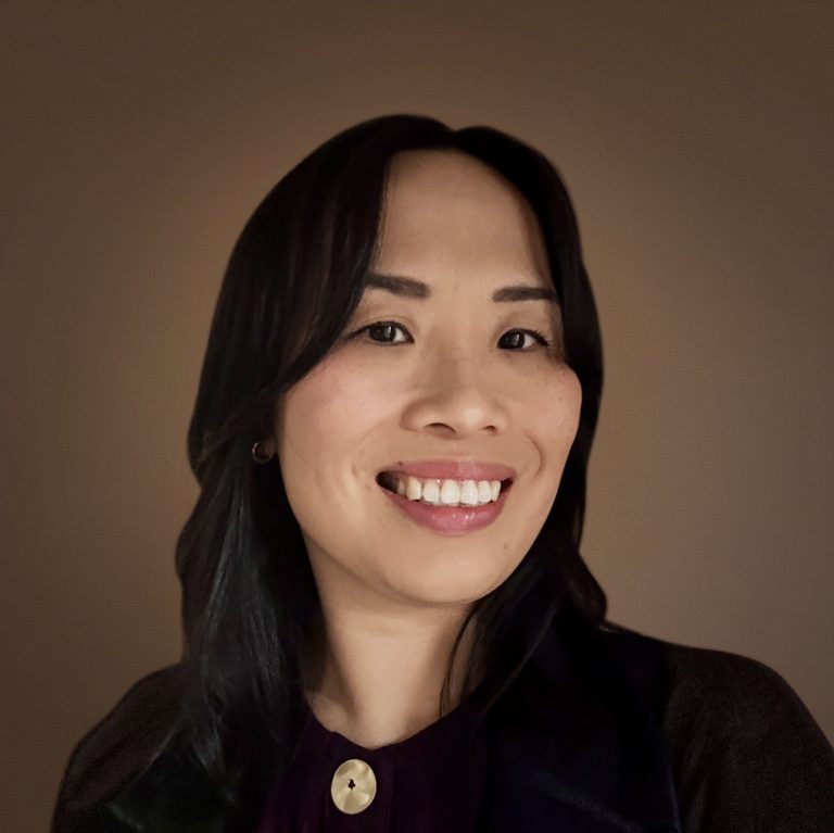 Woman with long dark hair smiles at the camera, wearing a dark top with a round button detail, against a plain brown background.