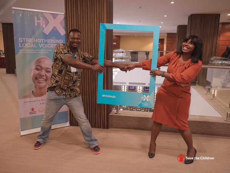Two smiling people hold a blue rectangular frame together indoors at the Humanitarian Xchange Abuja event. A banner and Save the Children logo are visible in the background. They appear joyful and are posing playfully for the photo.