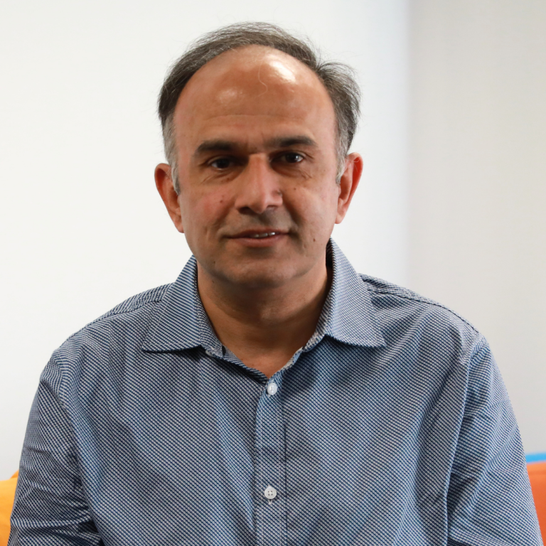 A man with short dark hair and a receding hairline, wearing a blue patterned button-up shirt, is sitting and smiling slightly in front of a plain light background.