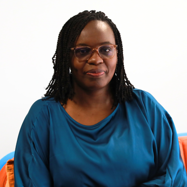 A woman with braided hair and glasses, wearing a blue top, sits on a colorful couch against a plain white background, looking at the camera with a calm expression.