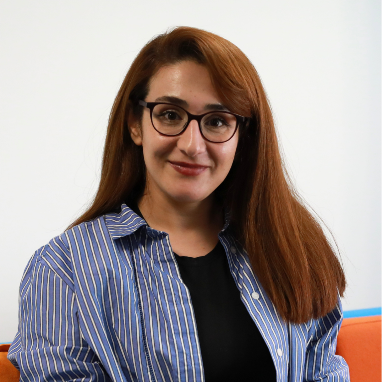 A woman with long reddish-brown hair and glasses smiles at the camera. She is wearing a blue and white striped shirt over a black top and is seated against a plain white background.