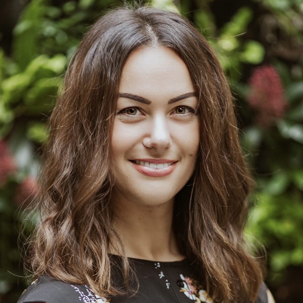 A woman with wavy brown hair and a floral black top smiles in front of a background of green plants and flowers.