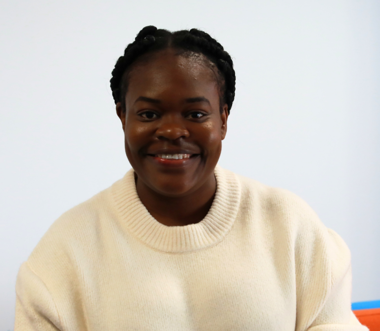 A person with braided hair is smiling and wearing a cream-colored sweater, seated against a plain white background.