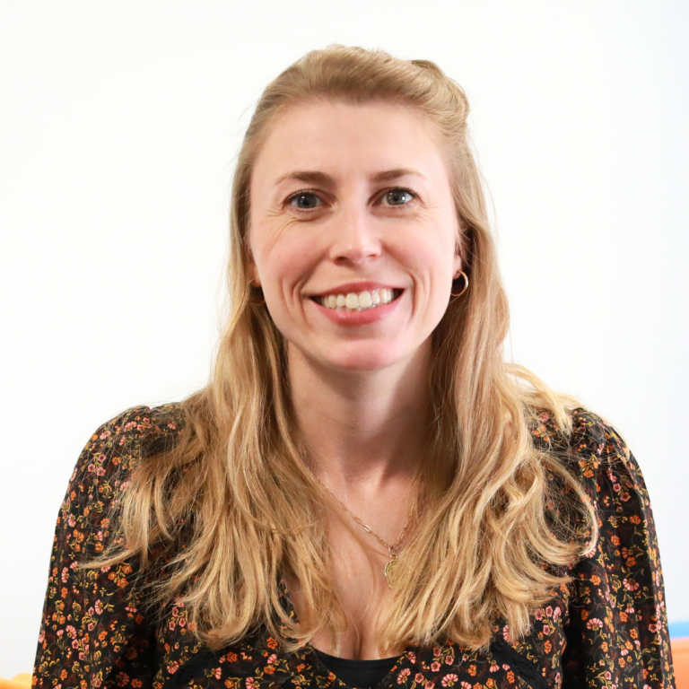 Woman with long blonde hair wearing a black floral top, smiling at the camera in front of a plain white background.