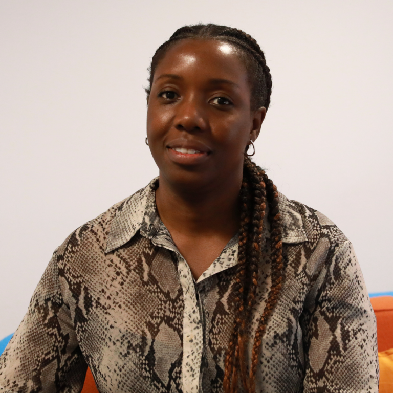 A woman with braided hair, wearing a patterned button-up shirt, sits indoors against a plain background, looking at the camera with a slight smile.