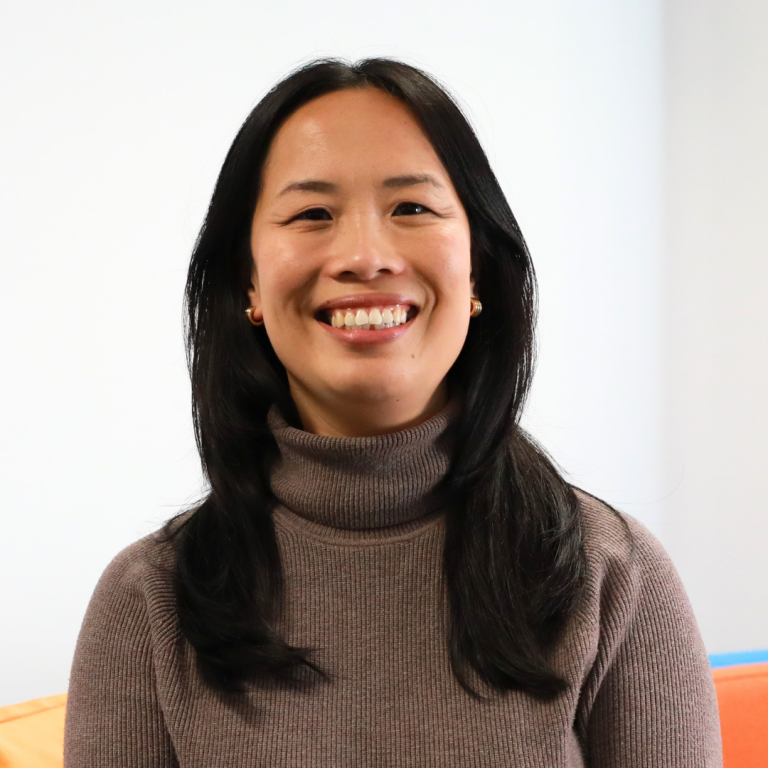 A woman with long dark hair smiles at the camera. She is wearing a brown turtleneck sweater and is seated indoors against a plain white background with a hint of an orange chair.