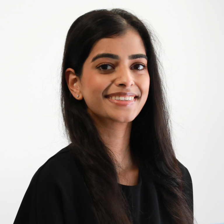 A woman with long dark hair and a black top smiles at the camera in front of a plain white background.