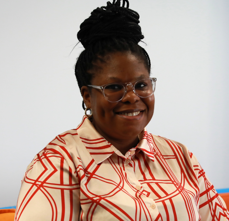 A woman with braided hair in a bun, wearing glasses and a patterned red and white shirt, sits smiling on an orange couch against a plain background.
