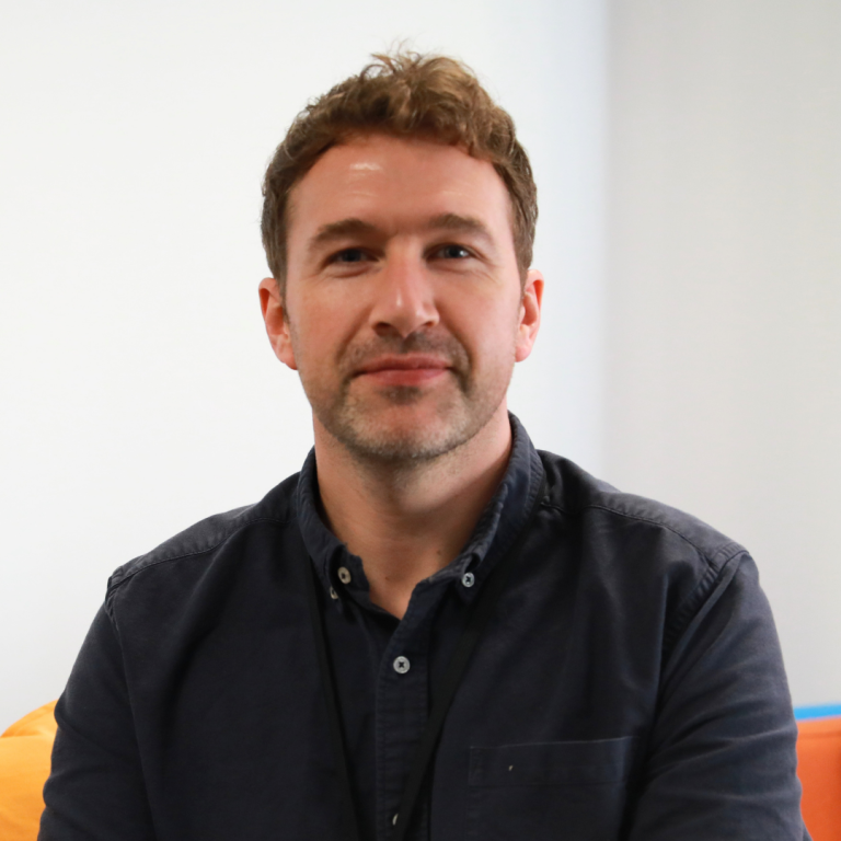 A man with short, light brown hair and a trimmed beard is sitting and smiling slightly. He is wearing a dark buttoned shirt and a lanyard. The background is simple, with light walls and part of an orange chair visible.