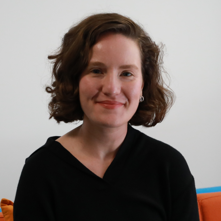 A person with short, curly brown hair and a black top smiles while sitting on a couch with orange and blue cushions, against a plain white background.