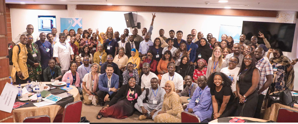 A large, diverse group of attendees pose for a group photo in a conference room. Some are standing, others sitting in chairs or on the floor. Many wear name tags and business-casual attire. Several individuals are smiling or waving to the camera.