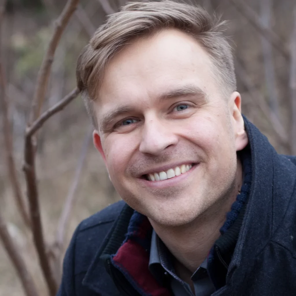 A man with light brown hair and blue eyes smiles warmly at the camera while outdoors, wearing a dark jacket. Leafless branches and a blurred natural background are visible behind him.