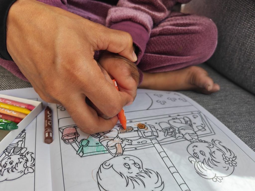 An adult helps a child color a drawing in a coloring book with crayons. Both hands hold an orange crayon, and only their hands and the childs legs are visible.