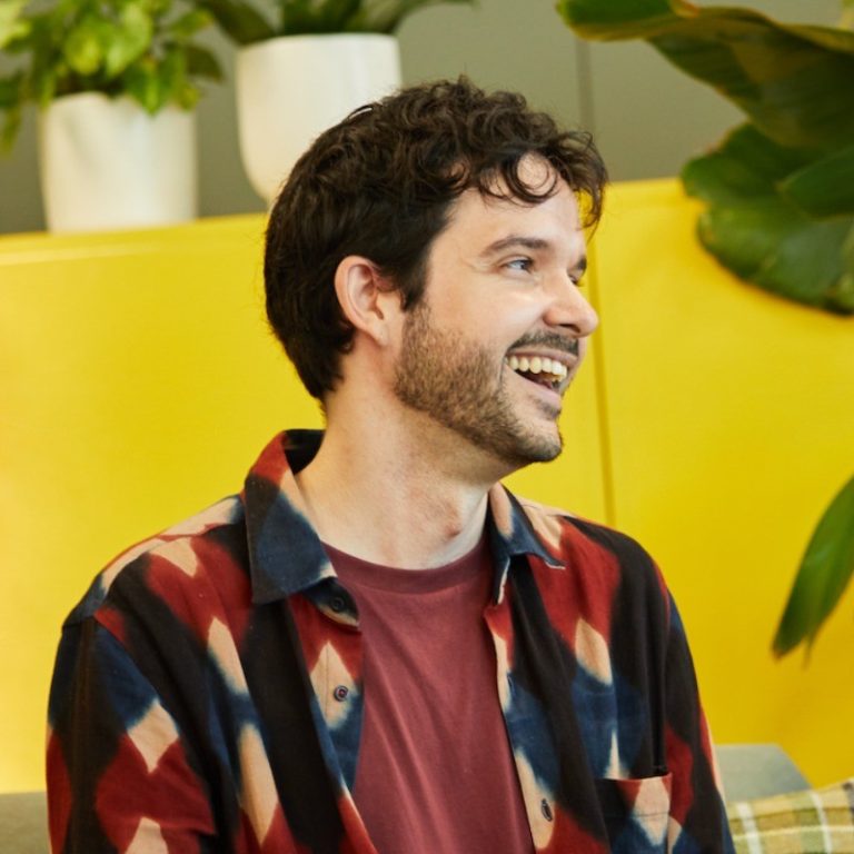 A man with dark curly hair and a beard smiles while looking to the side. He wears a patterned shirt over a maroon t-shirt. Set against green plants and a bright yellow wall, he embodies the spirit of permaculture and regenerative humanitarian response, reflecting a lifestyle attuned to sustainability and positive change.