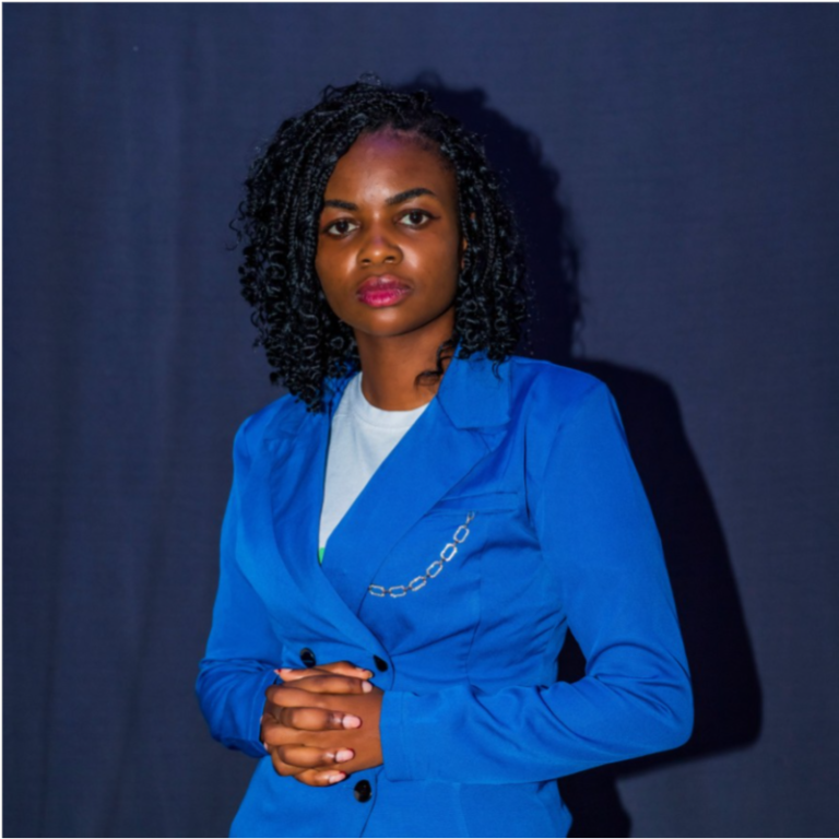 A woman with curly black hair wearing a bright blue blazer stands against a dark background, looking confidently at the camera with her hands clasped together, embodying the forward-thinking spirit of permaculture and regenerative humanitarian response.