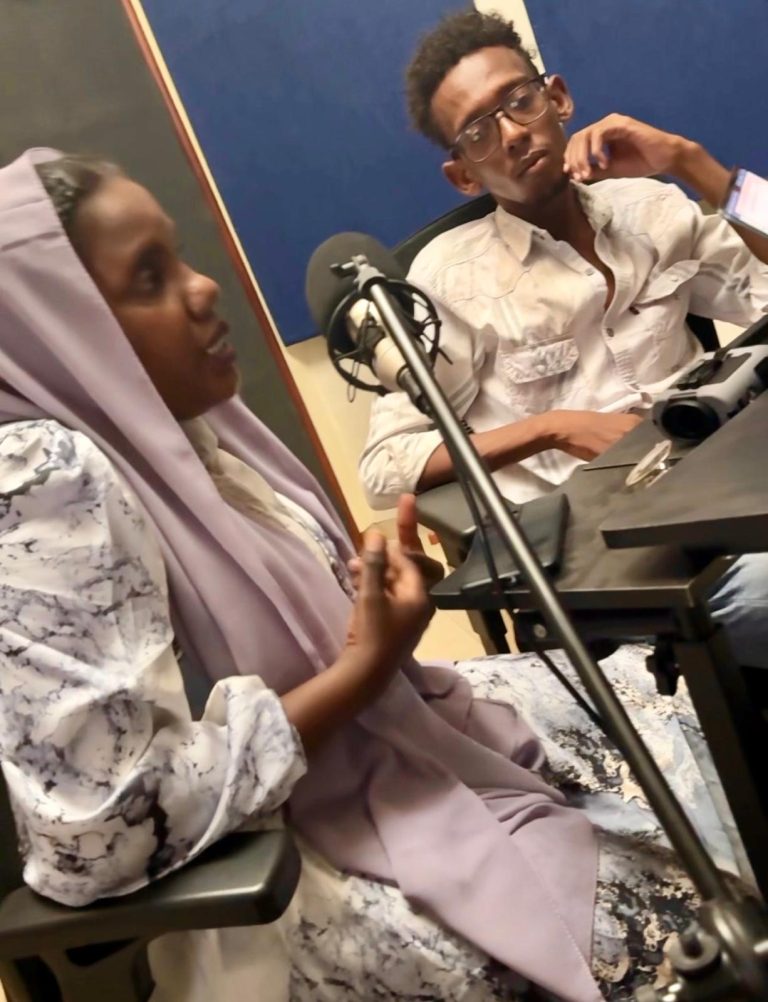 Two young people sit at a table in a recording studio. A girl in a patterned dress and light purple headscarf speaks into a microphone, while a boy in glasses listens attentively.