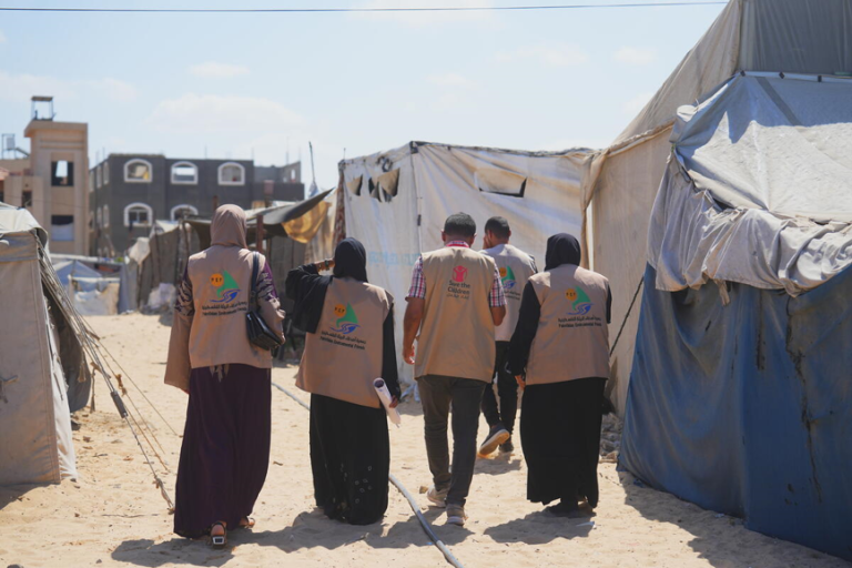 Four aid workers in tan vests walk through a sandy area lined with tents, possibly in a refugee or disaster relief camp, with buildings visible in the background under a clear sky.