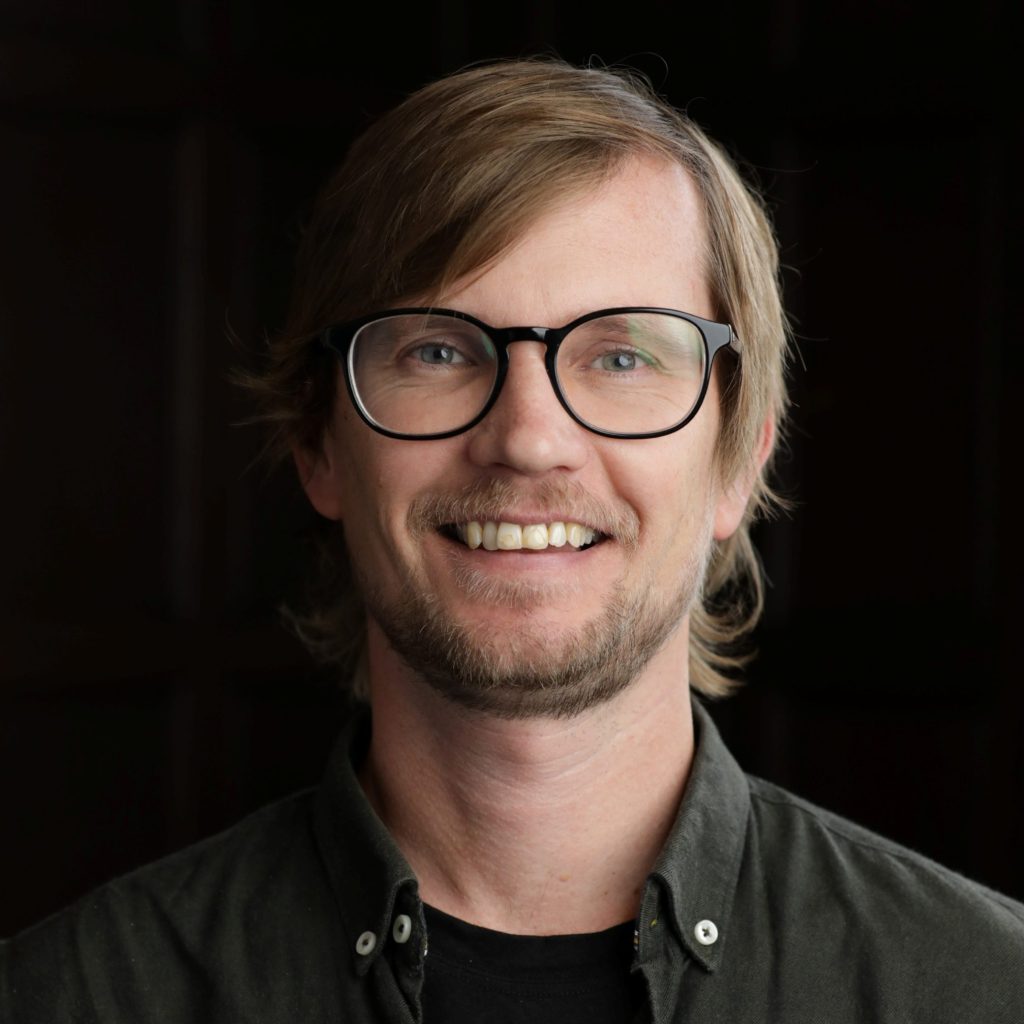 A smiling man with light brown hair, glasses, and a short beard wears a dark button-up shirt over a black t-shirt, standing in front of a dark, blurred background.