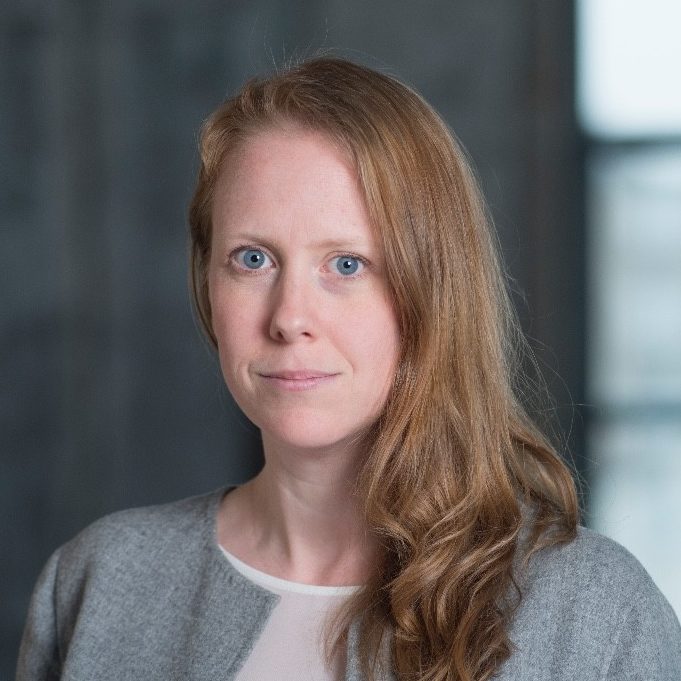 A woman with long, light brown hair and blue eyes wears a grey blazer over a white top, standing in front of a blurred dark background.