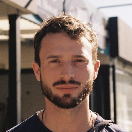 A man with short, curly brown hair and a beard stands outdoors, looking at the camera. The background is slightly blurred, showing building structures and sunlight.