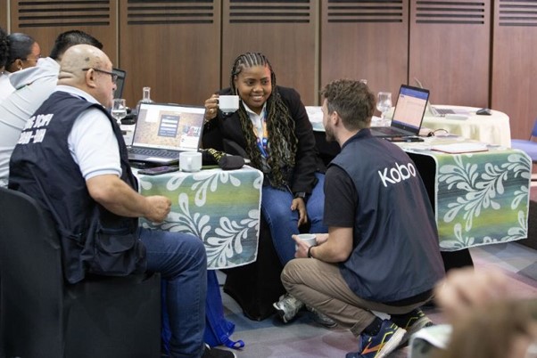 Three people in vests sit and kneel around a table with laptops, engaged in discussion at an indoor event. The table is covered with a green and white floral-patterned cloth.