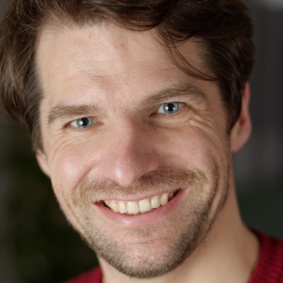 A man with light skin, brown hair, and blue eyes smiles at the camera. He has some facial hair and is wearing a red top. The background is blurred, hinting at a setting dedicated to humanitarian digital learning.