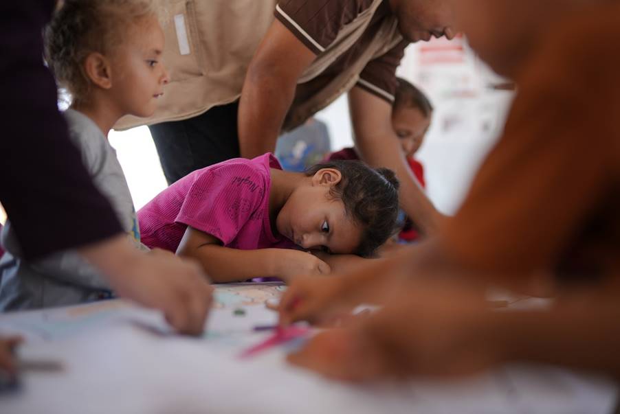 A group of children and an adult sit around a table working on a paper craft project together.